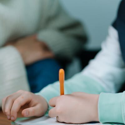 Close-up of a child's hands writing with an orange pen on a book indoors.